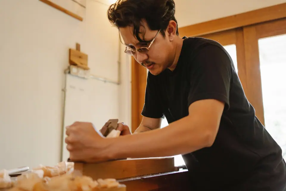 A craftsman planing a teak board at a bench, with wood shavings on the bench