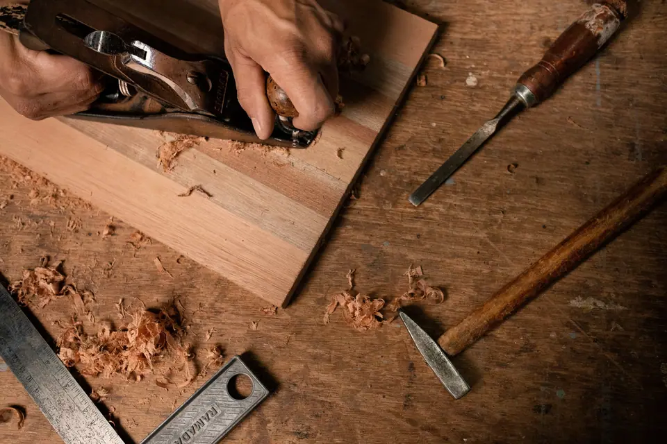 Hands planing a teak board beside a chisel and a square